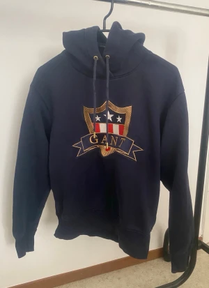 Marinblå hoodie från GANT med emblem - Snygg marinblå hoodie från GANT med broderat emblem på bröstet i rött, vitt och guld. Klassisk huva med dragsko och ribbade muddar. Perfekt för en avslappnad och stilren look.