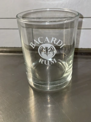 Bacardi Rum glas - Säljer ett genomskinligt glas med Bacardi Rum-logga tryckt i vitt. Glaset är cylinderformat och passar perfekt för drinkar eller som samlarobjekt för Bacardi-fans.