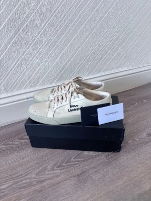 Saint Laurent vita canvas sneakers - Snygga vita sneakers från Saint Laurent i canvas med handskriven logga på sidan. Skorna har lågt skaft, vita snören och en klassisk gummisula. Perfekta för en clean och avslappnad stil.