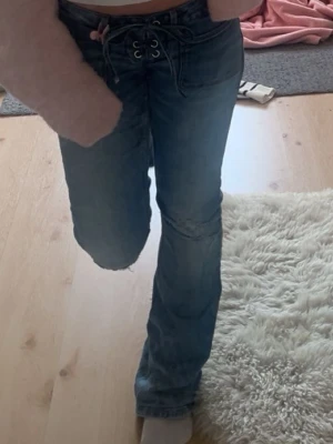 Blå bootcut jeans med snörning - Säljer ett par blå bootcut jeans med cool snörning framtill i midjan och slitningar på låret. Jeansen har en lätt tvättad look och är tillverkade i denim. Perfekt för dig som gillar trendiga detaljer och en avslappnad stil.