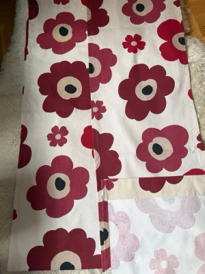 Marimekko Unikko tyg i rött och vitt - Säljer ett tygstycke från Marimekko med det klassiska Unikko-mönstret i rött, rosa och vitt. Tyget har stora blommor och är gjort i bomull. Perfekt för att sy egna inredningsdetaljer eller använda som duk.Mått 40 cm * 209 cm på varje längd.