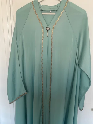 Ljusblå/turkås långklänning med gulddetaljer - Elegant ljusblå/turkås långklänning med guldfärgade broderade detaljer längs kanterna och en dekorativ hjärtformad brosch framtill. Klänningen har vida ärmar och ett mjukt, flödande material som ger en lyxig känsla. Perfekt för speciella tillfällen. PRIS GÅR ATT DISKUTERAS! 