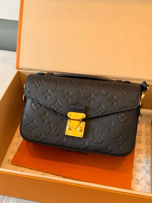 Svart Louis Vuitton handväska i läder - Snygg svart handväska från Louis Vuitton i präglat läder med klassiskt monogrammönster. Väskan har guldfärgat lås och detaljer samt ett stilrent, kompakt format. Perfekt accessoar för att lyfta din outfit.