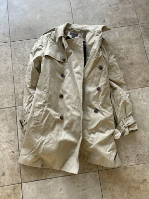 Beige klassisk trenchcoat - Stilren beige trenchcoat med dubbelknäppning, axelklaffar och bälte vid ärmsluten. Jackan har två snedställda fickor och klassisk krage. Perfekt för dig som vill ha en tidlös och snygg look. Materialet är troligtvis bomull eller bomullsblandning.