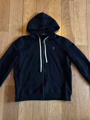 Svart hoodie från Polo Ralph Lauren - Snygg svart hoodie med dragkedja från Polo Ralph Lauren. Klassisk design med huva och vita snören, samt kängurufickor framtill. Liten röd logga broderad på bröstet. Perfekt för en avslappnad och stilren look.