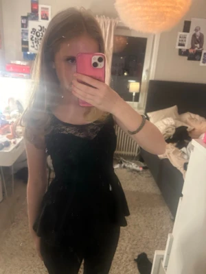 Svart peplum topp med spetsdetalj - Snygg svart peplum topp med spets upptill och rund halsringning. Toppen har korta ärmar och markerad midja som ger en feminin siluett. Perfekt att styla med jeans eller kjol för en trendig look.