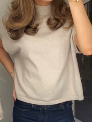 Beige stickad topp med krage - Snygg beige stickad topp med korta ärmar och ribbad krage. Perfekt att styla med jeans eller kjol för en clean och trendig look. Materialet ser mjukt och bekvämt ut, och modellen har en avslappnad passform.