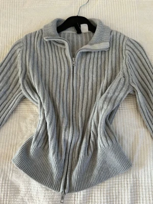 Ljusgrå stickad ziptröja - Snygg ljusgrå ribbstickad tröja med dragkedja framtill och hög krage. Tröjan har långa ärmar och en figurnära passform som ger en modern look.