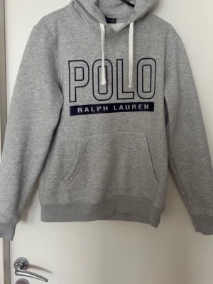 Grå hoodie från Polo Ralph Lauren - Snygg grå hoodie från Polo Ralph Lauren med stor logga framtill i mörkblått och vita dragsnören i huvan. Klassisk känguruficka och ribbade muddar. Tillverkad i mjuk bomullsblandning som är perfekt för chill dagar. Storlek m men passar även s