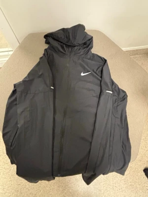 Svart Nike vindjacka med huva - Snygg svart vindjacka från Nike med dragkedja framtill och huva. Jackan har två sidofickor, diskreta reflexdetaljer och den klassiska Nike-loggan på bröstet. Tillverkad i lätt och vindtätt material, perfekt för blåsiga dagar.