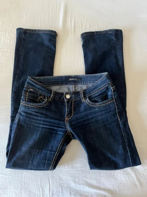 Mörkblå lågmidjade utsvängda jeans från Just F - Mörkblå lågmidjade utsvängda jeans från Just F med kontrastsömmar och klassisk femficksdesign. Jeansen har låg midja och bootcut passform, perfekta till en avslappnad streetstil. Materialet är denim med stretch för extra komfort. Skador vid knapp (syns på bild 5)