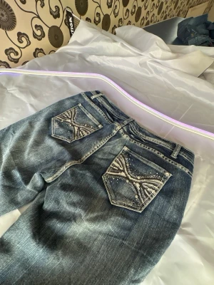 Blå bootcut jeans med blingade fickor Y2k Rosalina jeans - Säljer ett par blå bootcut jeans med snygga slitningar och dekorerade bakfickor med bling och broderier. Jeansen har klassisk femficksmodell och normal midja. Perfekta för dig som vill sticka ut lite extra med detaljer på fickorna.