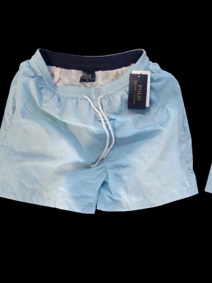 Ljusblå badbyxor från Polo Ralph Lauren - Snygga ljusblå badbyxor från Polo Ralph Lauren med elastisk midja och vit dragsko. Klassisk modell med meshfoder och bakficka med lock. Perfekta för stranden eller poolen, tillverkade i snabbtorkande material.
