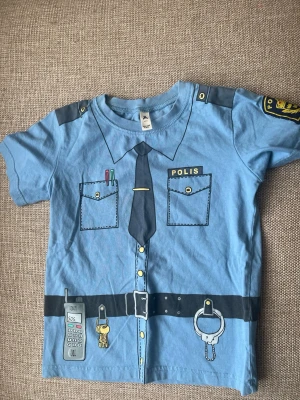 Blå polis t-shirt från Åhléns - Rolig blå t-shirt från Åhléns med tryck som föreställer en polisuniform. Trycket har detaljer som slips, fickor, polisbricka, radio, handbojor och nycklar. Perfekt för maskerad eller lek. Tillverkad i mjuk bomull.