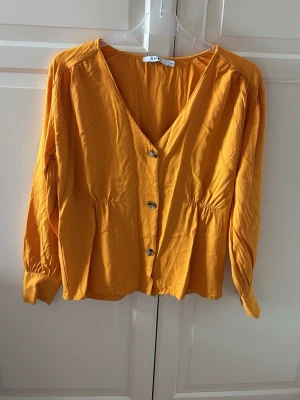 Gul blus med knappar från NA-KD - Trendig gul blus från NA-KD med v-ringning och dekorativa knappar framtill. Blusen har lång ärm, lätt rynk vid midjan och är tillverkad i ett mjukt, tunt tyg som ger en avslappnad look. I fint skick. Storlek M. Färgen är senapsgul. 