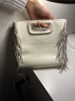 Maje handväska  - En fin vit mage väska i croco-mönstrat konstläder med fransar på sidorna och ett inbyggt handtag. Väskan har även en avtagbar axelrem för flexibel användning. Perfekt för dig som gillar trendiga detaljer och stilren design.
