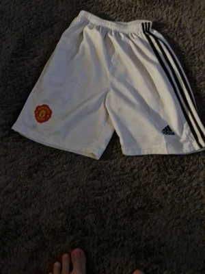 Vita Manchester United shorts Adidas - Säljer ett par vita Manchester United shorts från Adidas med svarta ränder på sidan och klubbens emblem broderat på ena benet. Perfekta för fotboll eller träning, med elastisk midja för skön passform.