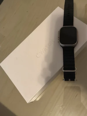 Apple Watch Ultra med svart armband - Snygg Apple Watch Ultra med svart silikonarmband och silverfärgat aluminiumhölje. Klockan har en fyrkantig form med rundade hörn och en stor, tydlig skärm. Perfekt för dig som vill ha koll på både träning och notiser direkt på handleden.