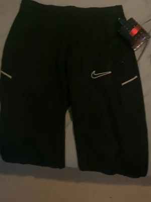 Svarta Nike mjukisbyxor med logga - Säljer ett par svarta mjukisbyxor från Nike med vit broderad Swoosh-logga och vita detaljer vid fickorna. Byxorna har resår i midjan och muddar vid bensluten. Perfekta för chill dagar eller träning.