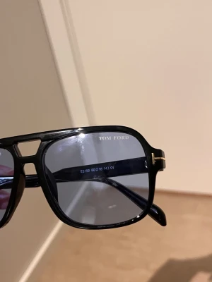 Svarta Tom Ford solglasögon - Snygga svarta solglasögon från Tom Ford med klassisk fyrkantig form och mörka glas. Bågarna har en diskret gulddetalj vid gångjärnen och är tillverkade i Italien. Perfekta för dig som vill ha en stilren och exklusiv look.