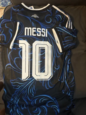Argentina Messi tröja VM 2026 - Snygg Argentina fotbollströja från Adidas med Messi och nummer 10 på ryggen. Tröjan är svart med blåa mönster, vita detaljer och officiella märken från FIFA och AFA. Korta ärmar, v-ringad och tillverkad i lätt funktionsmaterial.
