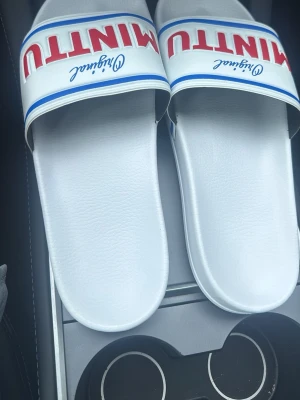 Vita Minttu slides med blå och röd text - Säljer ett par sprillans nya vita tofflor från Minttu.