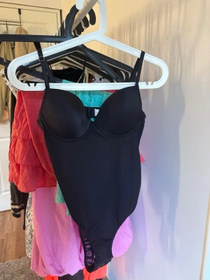 Svart body med bygel och tunna band - Snygg svart body med vadderade kupor och bygel, tunna justerbara axelband och slät finish. Perfekt för att skapa en stilren och enkel look. Materialet känns mjukt och stretchigt mot huden.
