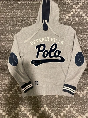 Grå hoodie Beverly Hills Polo Club - Snygg grå hoodie från Beverly Hills Polo Club med stor logga framtill. Mörkblå detaljer på armbågar och muddar, samt vita och blå ränder vid ärmslut och midja. Huva med dragsko och mjukt material, perfekt för en avslappnad stil. Mycket bra skick, storlek M.