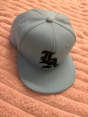 Ljusblå LA snapback keps - Snygg ljusblå snapback keps med svart LA-logga framtill. Klassisk platt skärm och justerbar baksida. Tillverkad i slitstark bomullsmix, perfekt för en streetwear-look.