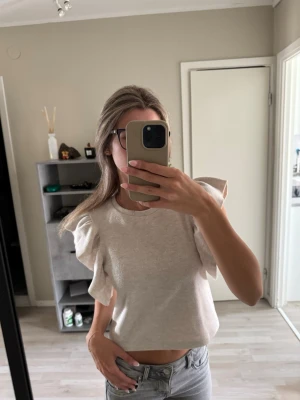 Beige topp med volangärmar - Trendig beige topp med korta volangärmar och croppad passform. Toppen har rund halsringning och är tillverkad i ett mjukt material som känns skönt mot huden. Kom till användning 2 gånger.