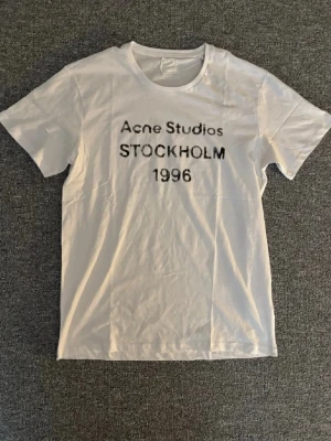 Vit Acne Studios t-shirt Stockholm - Vit t-shirt från Acne Studios med svart text 'STOCKHOLM 1996' på bröstet. Klassisk passform och rund halsringning, perfekt för en clean och stilren look. Tillverkad i mjuk bomull.