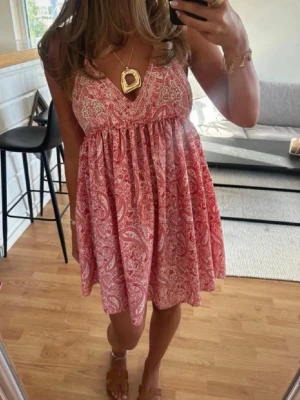  Trendig Rosa paisleymönstrad kortklänning - Säljer en söt kortklänning med paisleymönster i rosa och vitt. Klänningen har v-ringad hals, tunna axelband med knyt och ett luftigt fall. Perfekt för varma dagar och enkel att styla med smycken. Materialet känns lätt och bekvämt.