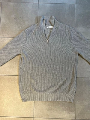 Grå stickad halvzip från Zara - Säljer en stilren grå stickad tröja från Zara med halv dragkedja och hög krage. Tröjan har ribbstickad struktur, långa ärmar och bred mudd vid ärmslut och nederkant. Perfekt för lager på lager och enkel att matcha.