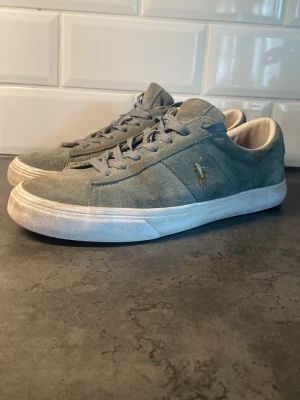 Grå gröna mocka sneakers från Polo Ralph Lauren - Pris diskuteras. Snygga grå sneakers i mocka från Polo Ralph Lauren med klassisk låg profil och vit sula. Skorna har grå skosnören och den ikoniska broderade loggan i guld på sidan. Perfekta för en clean och stilren look.