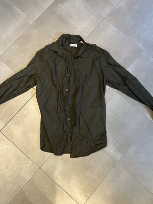 Mörkgrön skjorta från Jack&Jones - Stilren mörkgrön skjorta från jack&jones med klassisk krage och knäppning framtill. Skjortan har lång ärm och är tillverkad i ett tunt, mjukt material som känns skönt mot huden. Perfekt för dig som gillar en enkel men snygg look.