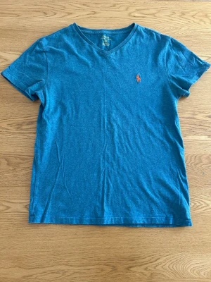 Blå v-ringad t-shirt från Polo Ralph Lauren - Snygg blå t-shirt från Polo Ralph Lauren med v-ringning och klassisk orange logga broderad på bröstet. Tillverkad i mjuk bomull och har en normal passform. Perfekt till jeans eller shorts för en clean och enkel stil.