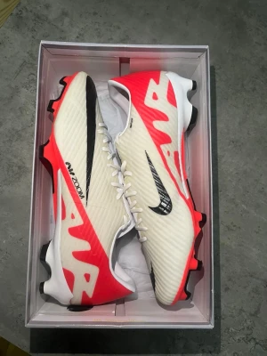 Nike Air Zoom Mercurial fotbollsskor - Säljer ett par Nike Air Zoom Mercurial fotbollsskor i vitt med svarta och neonröda detaljer. Skorna har en låg profil, snörning och mönstrad yttersula för bra grepp på planen. Tillverkade i syntetmaterial för lätt vikt och snabbhet. Storlek 44 men passar till 43 eftersom Nike har tighta skor.