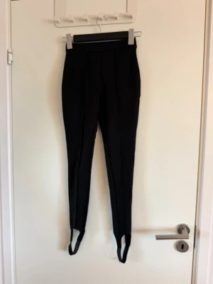 Svarta stretchiga leggings med fotstrap - Säljer ett par svarta leggings med fotstrap och hög midja. De har en smal passform och söm framtill som ger en snygg siluett. Perfekta till sneakers eller boots. Materialet är stretchigt och bekvämt. Aldrig använd endast testad. Köpt på vinted.