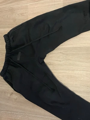 Svarta mjukisbyxor från essentials  - Säljer ett par svarta mjukisbyxor från essentials med diskret logga på benet. Byxorna har elastisk midja med dragsko och muddar vid benslut. Perfekta för chill eller träning, tillverkade i mjukt bomullsmaterial som är riktigt skönt mot huden.