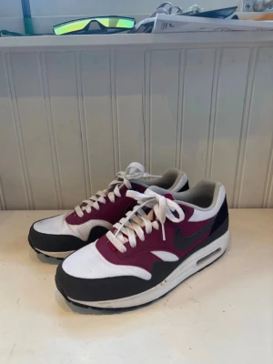 Nike Air Max 1 vinröd/vit/svart - Nike Air Max 1 sneakers i vinrött, vitt och svart med klassisk swoosh på sidan. Skorna har lågt skaft, vita snören och synlig Air-sula. Ovandelen är i textil och mocka för en schysst retrostil. Perfekta för dig som gillar streetwear och sneakerskultur. Kan sälja billigare vid snabb affär 
