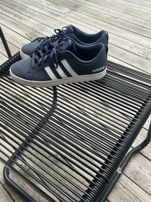 Mörkblå Adidas sneakers med vita ränder - Snygga mörkblå sneakers från Adidas med klassiska vita stripes på sidorna och vit sula. Skorna har låga skaft och är tillverkade i textil och syntetmaterial. Perfekta för dig som gillar en clean och sportig look. Använda 1 gång, pris kan diskuteras!