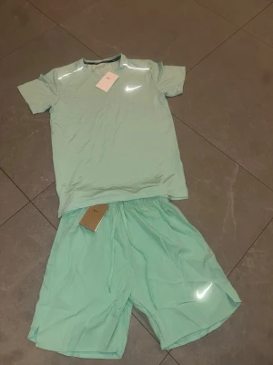 Mintgrönt träningsset från Nike - Säljer ett fräscht mintgrönt träningsset från Nike med t-shirt och shorts. Båda plaggen har den klassiska Nike-loggan och är gjorda i ett lätt och snabbtorkande material, perfekt för träning eller gym. Snygg och sportig look!