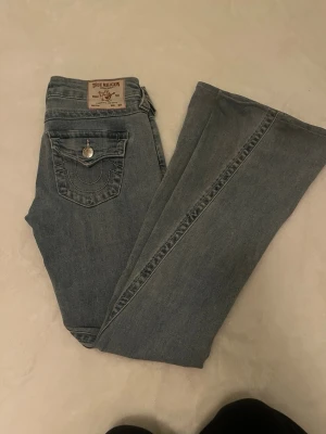 True Religion ljusblå jeans - Ljusblå jeans från True Religion med klassiska bakfickor och dekorativa sömmar. Jeansen har raka ben och silverfärgade knappar på fickorna. Materialet är denim och passformen är avslappnad. Perfekt för dig som gillar streetstyle och ikoniska märken.  (uppsydda)