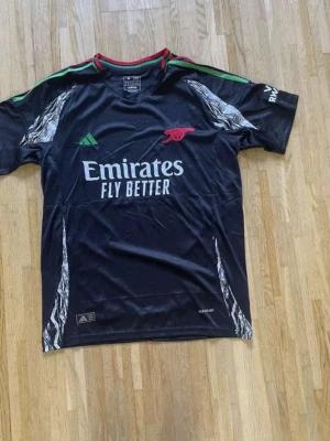 Arsenal svart matchtröja Adidas - Snygg svart Arsenal matchtröja från Adidas med gröna och röda detaljer på axlarna. Tröjan har trycket 'Emirates Fly Better' på bröstet och klubbens logga i rött. Tillverkad i lätt och ventilerande material, perfekt för träning eller match.