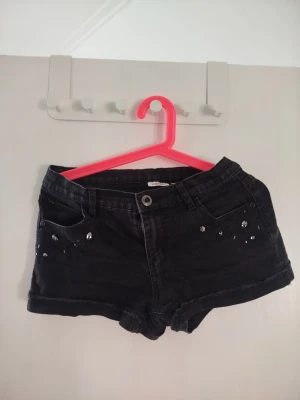 Svarta shorts med nitar och strass - Snygga svarta shorts med  dekorativa svarta stenar som glittrar på fickorna framtill även två små stenar på back fickorn. Perfekta för dig som vill ha en edgy och trendig look.