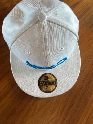 Vit New Era 59FIFTY keps med blå brodyr - Säljer en vit New Era 59FIFTY keps med platt skärm och blå broderad text framtill. Klassisk snapback-modell i bomullstyg med guldigt klistermärke kvar på skärmen. Perfekt för dig som gillar streetwear och vill sticka ut.