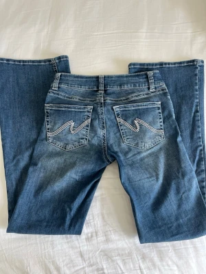 Blå bootcut jeans från Nelly - Säljer ett par blå bootcut jeans från Nelly med dubbla knappar i midjan och snygga detaljer på bakfickorna. Jeansen har klassisk femficksdesign och är gjorda i mjukt denimtyg. Perfekta för dig som gillar en avslappnad men trendig look.