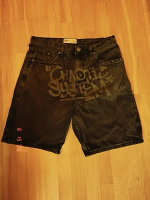 Svarta denimshorts med tryck - Unika svarta denimshorts från ISM med graffiti-inspirerat tryck 'Chaotic System' på framsidan och ett cirkelmönster på bakfickan. Klassisk femficksmodell med knappgylf och raka ben. Perfekta för dig som gillar streetwear och statement-plagg.