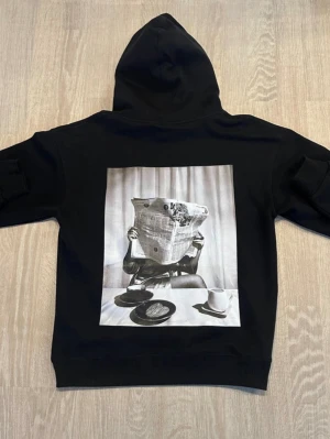 Svart hoodie med tidningsmotiv - Svart hoodie med stor huva och magficka. På ryggen finns ett stort svartvitt tryck av en person som läser tidningen vid ett frukostbord. Hoodien har en avslappnad passform och är tillverkad i mjukt bomullsmaterial. Perfekt för dig som gillar streetwear och unika prints.