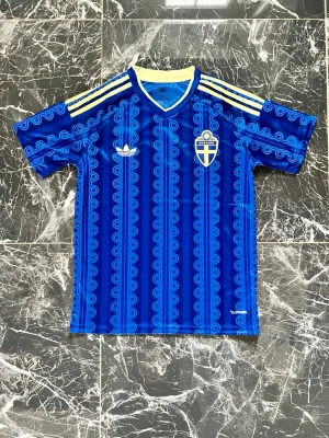 Blå Sverige fotbollströja Adidas - Säljer en blå fotbollströja från Adidas med Sveriges landslagsemblem på bröstet. Tröjan har gula detaljer, klassiska Adidas-ränder på axlarna och ett unikt mönster i olika blå nyanser. Perfekt för match eller träning.